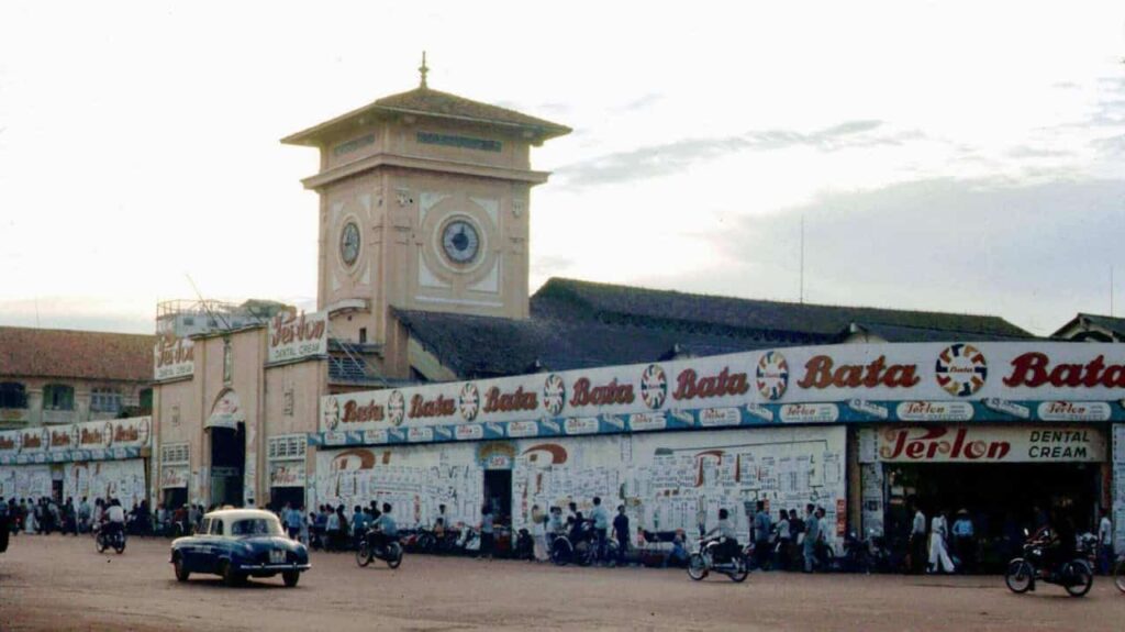 Historic Ben Thanh Market in Saigon, Vietnam, in 1967, showing its clock tower, storefronts, vintage cars, motorcycles, and busy streets.