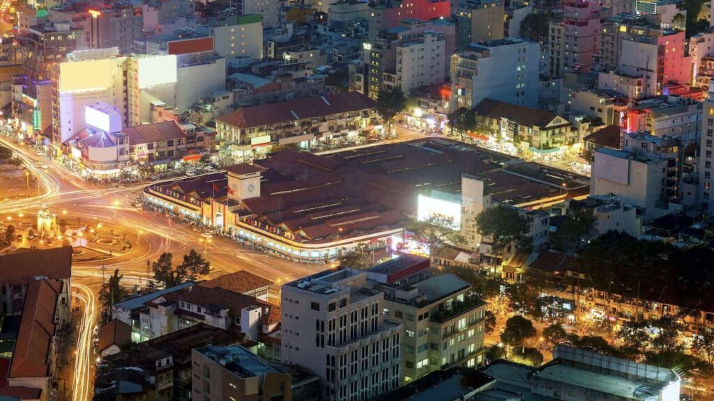 Aerial night view of Ben Thanh Market surrounded by brightly lit streets and buildings in central Ho Chi Minh City, Vietnam.