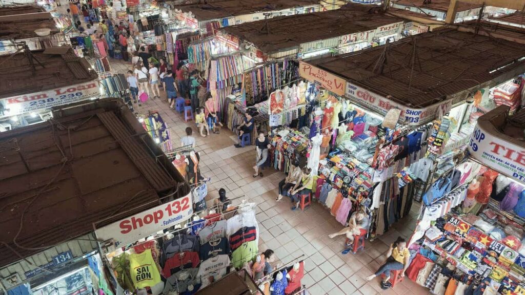 Busy interior of Ben Thanh Market in Ho Chi Minh City with colorful clothing, fabric, souvenirs, and local vendors.