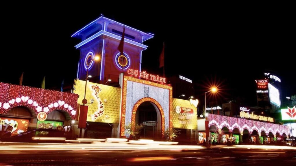 Ben Thanh Market illuminated at night with colorful lights, its iconic clock tower, and busy traffic in Ho Chi Minh City.